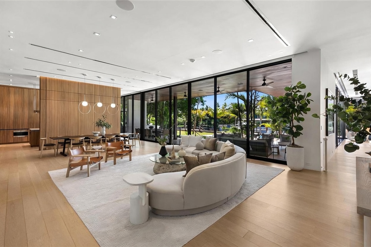 This expansive, open-concept living and dining area features light wood flooring and a neutral color palette that emphasizes a bright, airy atmosphere. A large, curved sofa sits on a textured rug, facing a wall of floor-to-ceiling glass that provides a seamless transition to the outdoor patio and lush greenery beyond. The space is accented by warm wood-paneled walls and modern pendant lighting, creating a sophisticated and inviting environment for entertaining.