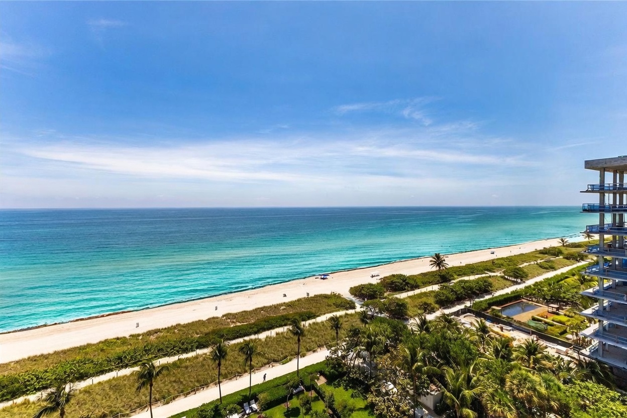This high-angle aerial view captures a stunning coastal panorama, showcasing a pristine sandy beach meeting the vibrant turquoise waters of the ocean. To the right, a portion of a modern high-rise building is visible, featuring balconies that overlook the lush green landscaping and walking paths below. The scene conveys a serene, luxurious beachside lifestyle with clear blue skies stretching across the horizon.
