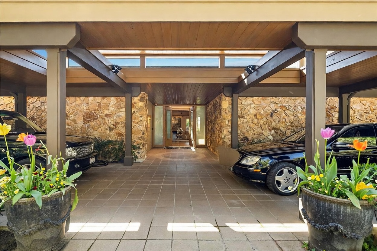 This grand, covered entryway features a porte-cochère design with dark wood beams and a tiled floor leading directly to the front doors. The walls are constructed from textured, multi-toned stone, creating a rustic yet sophisticated aesthetic. Two large planters with colorful tulips frame the entrance, while two luxury vehicles are parked on either side of the driveway, emphasizing the home's spacious and upscale arrival area.