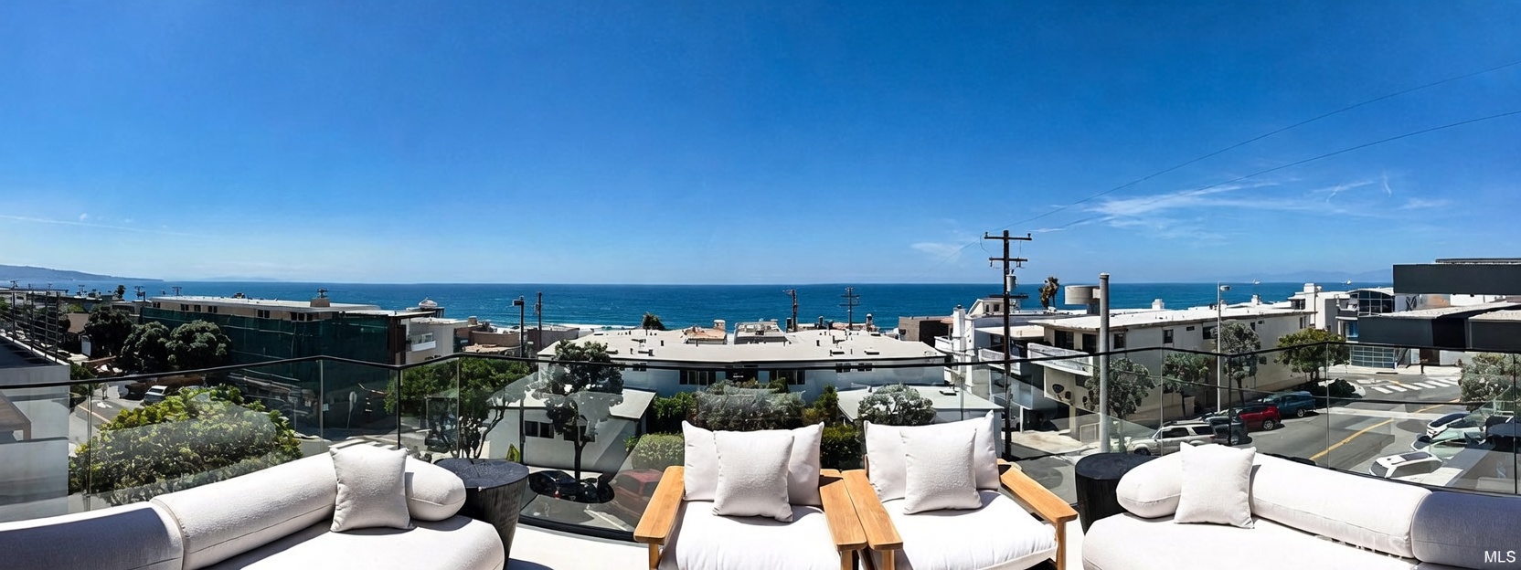 This expansive balcony offers a panoramic, unobstructed view of the Pacific Ocean under a clear blue sky. The space is furnished with comfortable, light-toned outdoor seating, including plush sofas and armchairs arranged to face the horizon. A sleek glass railing ensures the view remains the focal point, creating a seamless transition between the private terrace and the coastal landscape below.