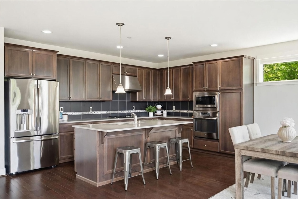 This spacious kitchen features dark wood cabinetry, stainless steel appliances, and a central island with seating for three. A dark tile backsplash contrasts with the light-toned countertops, while two pendant lights hang above the island to provide focused illumination. The open-concept design flows seamlessly into an adjacent dining area, creating a functional and inviting space for cooking and entertaining.