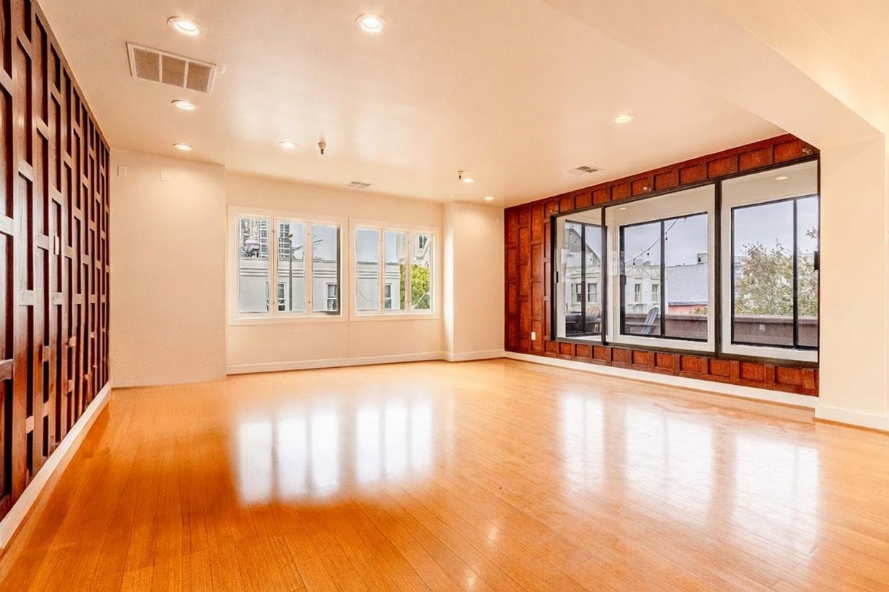This spacious living room features light wood flooring and a distinctive wall treatment with a dark wood grid pattern. Large windows on the left and a wide set of glass doors on the right provide ample natural light and views of the surrounding urban environment. The room has a clean, open layout with recessed lighting, creating a bright and versatile living space.