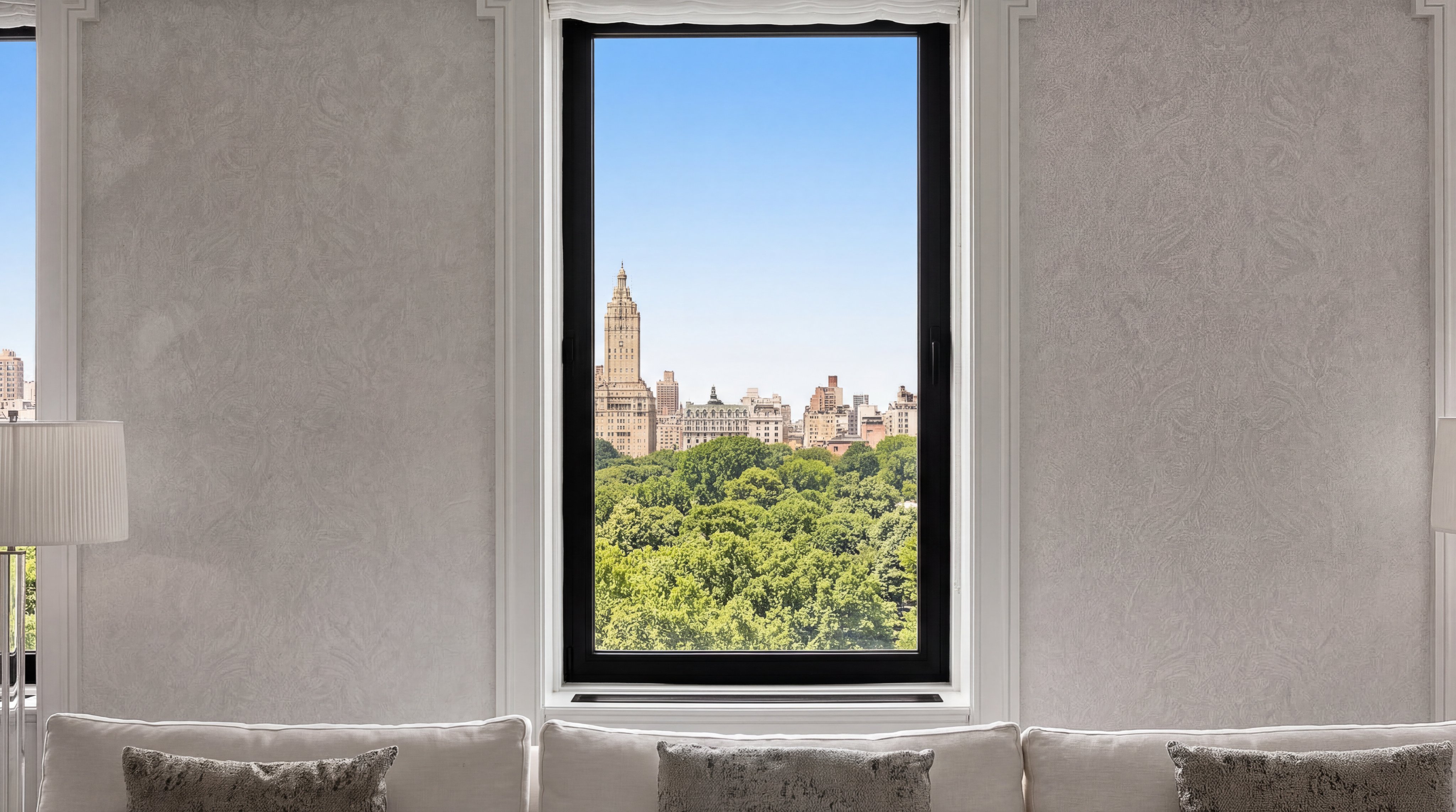 This interior shot highlights a bright living room space featuring a central, floor-to-ceiling window that frames a stunning, unobstructed view of a cityscape and lush parkland. The room is styled with elegant, neutral-toned furniture, including a plush sofa with soft throw pillows and a tall, minimalist floor lamp. The clean aesthetic and prime urban view suggest a high-end luxury property located in a major city like New York.