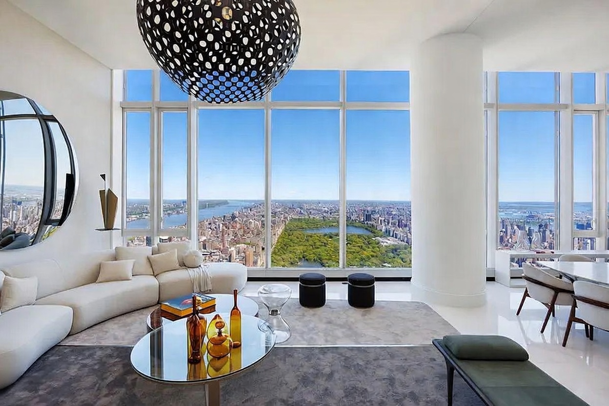 This sophisticated high-rise living room features an expansive, open-concept layout defined by floor-to-ceiling windows that offer a breathtaking, panoramic view of Central Park and the surrounding city skyline. The space is furnished with a plush, cream-colored sectional sofa, a modern glass-topped coffee table, and a striking, oversized spherical pendant light that serves as a focal point. The minimalist aesthetic is complemented by a neutral color palette, polished flooring, and a sleek, contemporary design that emphasizes the dramatic urban backdrop.