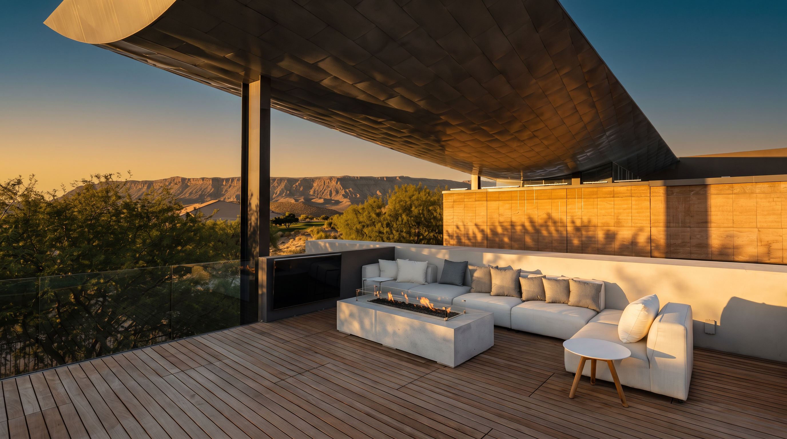 This expansive, modern outdoor deck features warm-toned wood plank flooring and a sleek, cantilevered metal roof structure that provides partial shade. The space is furnished with a contemporary white sectional sofa, a rectangular fire pit, and a small side table, all set against a backdrop of desert landscape and distant mountains. A glass railing maintains an unobstructed view, creating a seamless transition between the elevated living area and the natural surroundings.