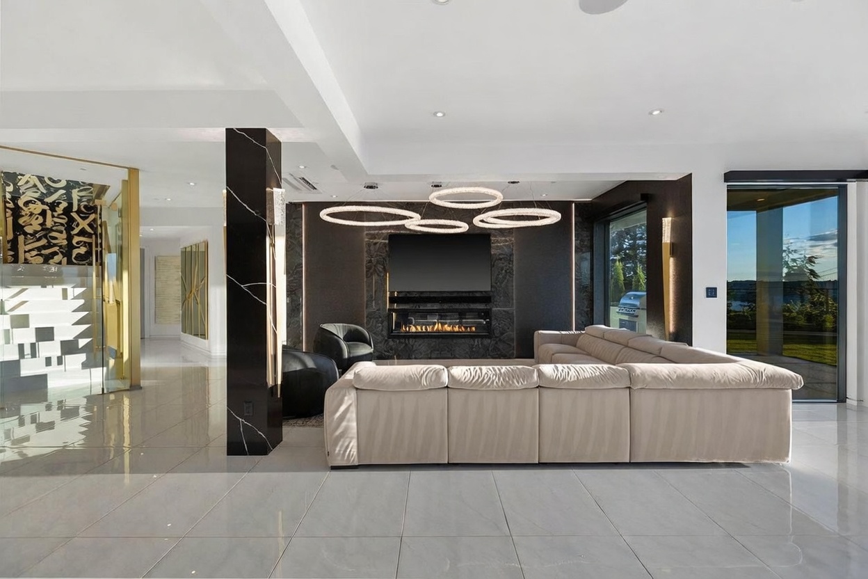 This modern living room features a large, light-colored sectional sofa facing a dark stone-clad chimney breast with an integrated fireplace and television. The space is illuminated by unique, circular pendant light fixtures and features polished tile flooring throughout. A large glass wall provides a view of the outdoor area, creating a seamless transition between the indoor living space and the exterior.