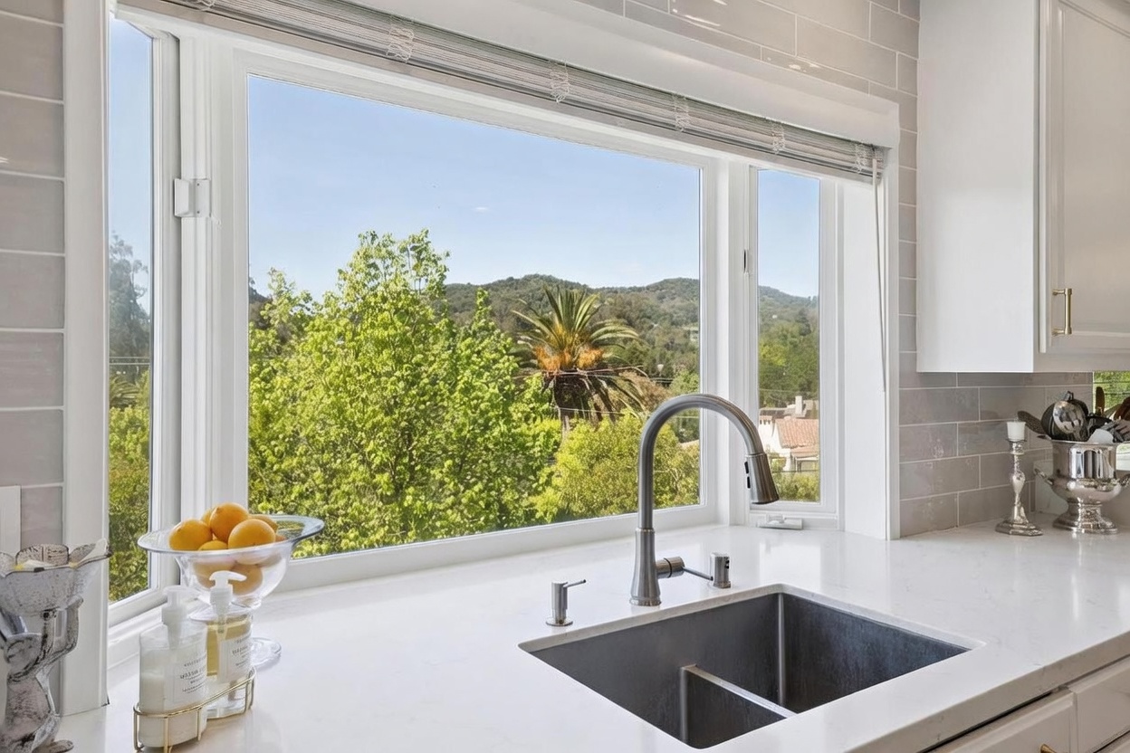 This bright kitchen features a clean, modern aesthetic with white cabinetry and light-toned stone countertops. A large, multi-pane window above the stainless steel sink provides a scenic, elevated view of lush green trees and distant hills. The space is accented by a neutral-toned subway tile backsplash and a bowl of fresh lemons, creating a welcoming and airy atmosphere.