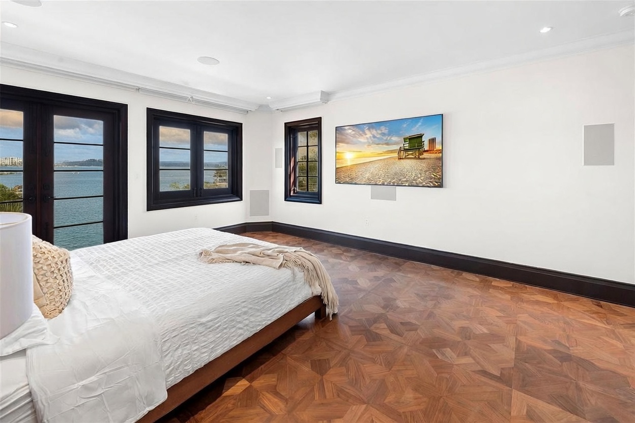 This spacious primary bedroom features rich, patterned wood flooring and crisp white walls, creating a bright and airy atmosphere. A large television is mounted on the wall, and the room includes a set of dark-framed glass doors that open to a scenic waterfront view. The space is anchored by a bed with neutral-toned bedding, offering a serene and sophisticated retreat.