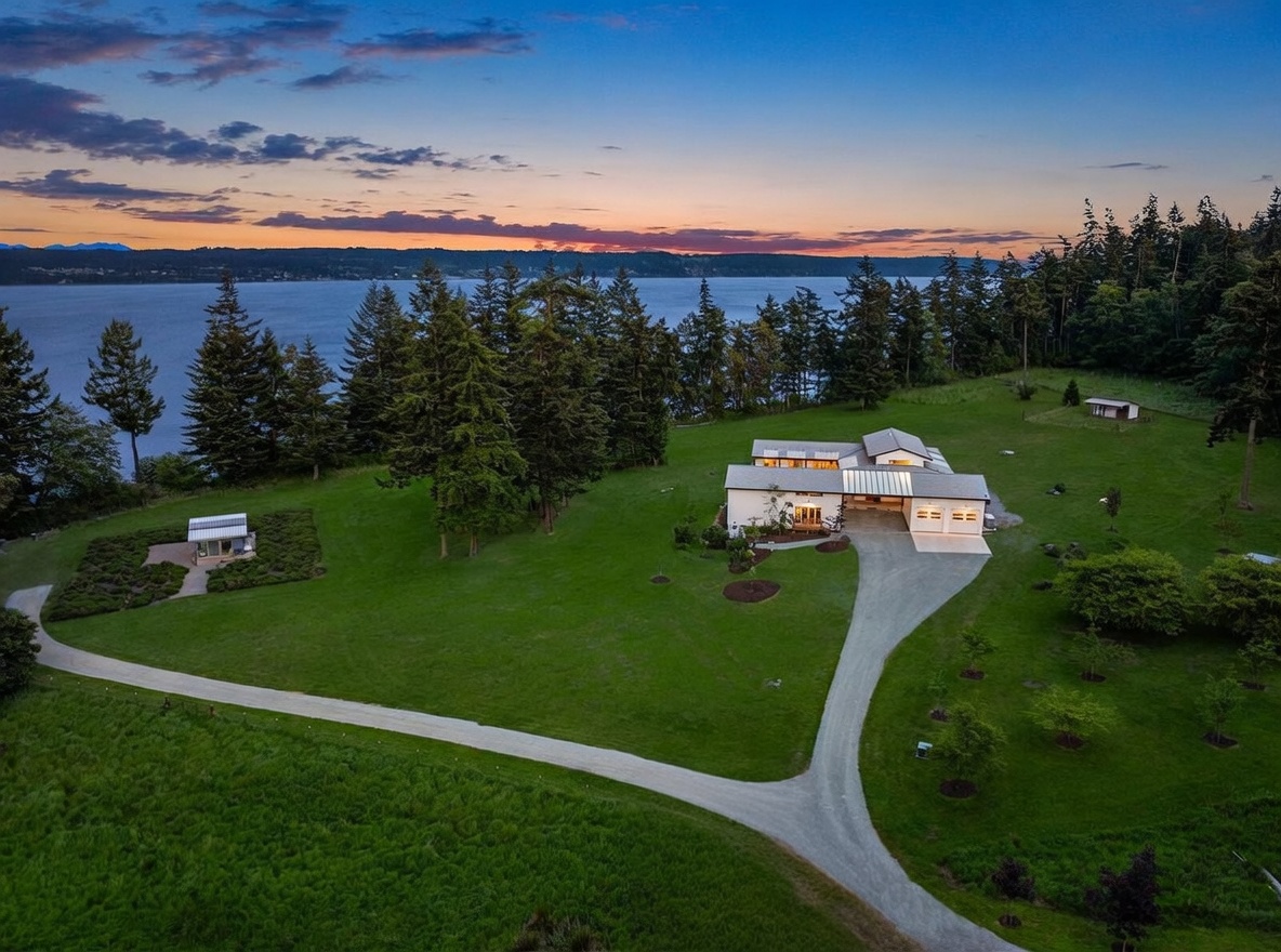 This high-angle aerial view captures a modern, single-story residence situated on a sprawling, lush green property overlooking a vast body of water at sunset. The home features a multi-gabled roofline, a gravel driveway leading to a multi-car garage, and a separate small structure set within a landscaped garden area. The expansive lawn is framed by mature evergreen trees, creating a serene and private atmosphere with a dramatic, colorful sky in the background.