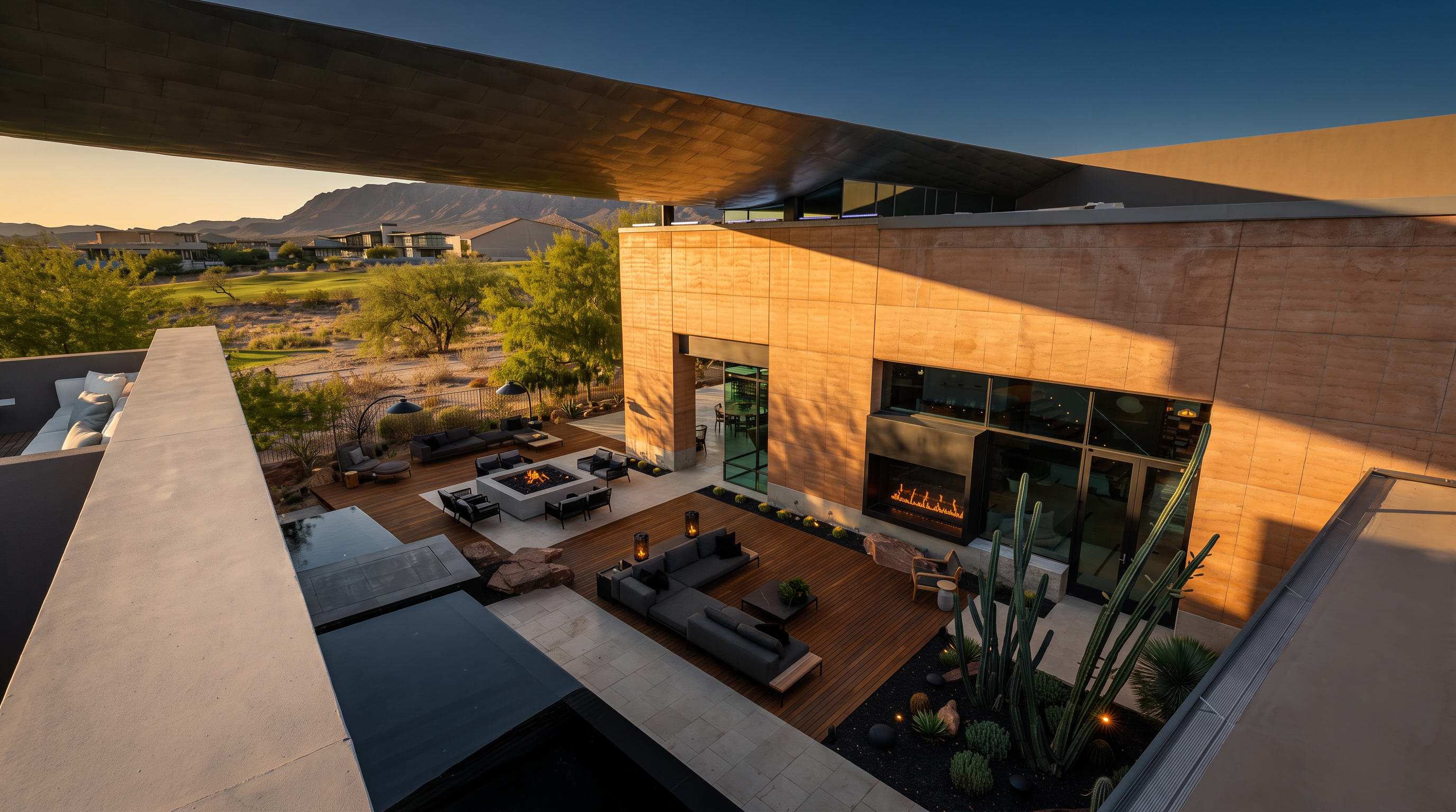 This high-angle perspective captures a sophisticated outdoor living space featuring a multi-level deck with dark wood planking and a built-in fire pit area. The architecture showcases a modern aesthetic with a prominent, cantilevered roof overhang and a large, linear fireplace integrated into the exterior wall. A desert-inspired landscape with cacti and dark gravel accents complements the clean lines of the structure, while the background reveals a scenic view of a golf course and distant mountains at sunset.
