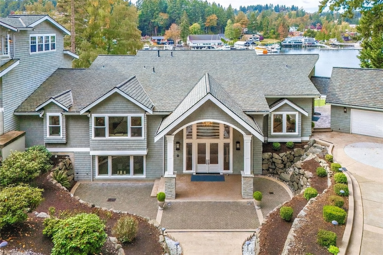 This elevated aerial perspective captures the front facade of a multi-level home featuring gray shingle siding and a prominent covered entryway with white columns. The property includes a herringbone-patterned stone patio, tiered landscaping with stone retaining walls, and a view of a nearby marina and forested hillside. The home's architectural design emphasizes a blend of traditional and coastal elements, creating an inviting and upscale curb appeal.