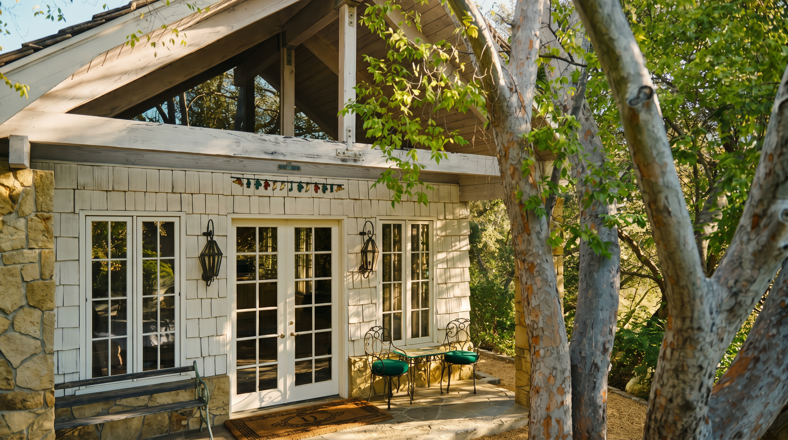 This charming cottage-style exterior features a stone-paved patio area shaded by mature trees, creating a serene and inviting atmosphere. The structure showcases white shingle siding, a prominent A-frame roofline with exposed timber framing, and classic French doors that open onto the patio. A wrought-iron bench and bistro set provide a cozy seating area, emphasizing the home's rustic and peaceful aesthetic.