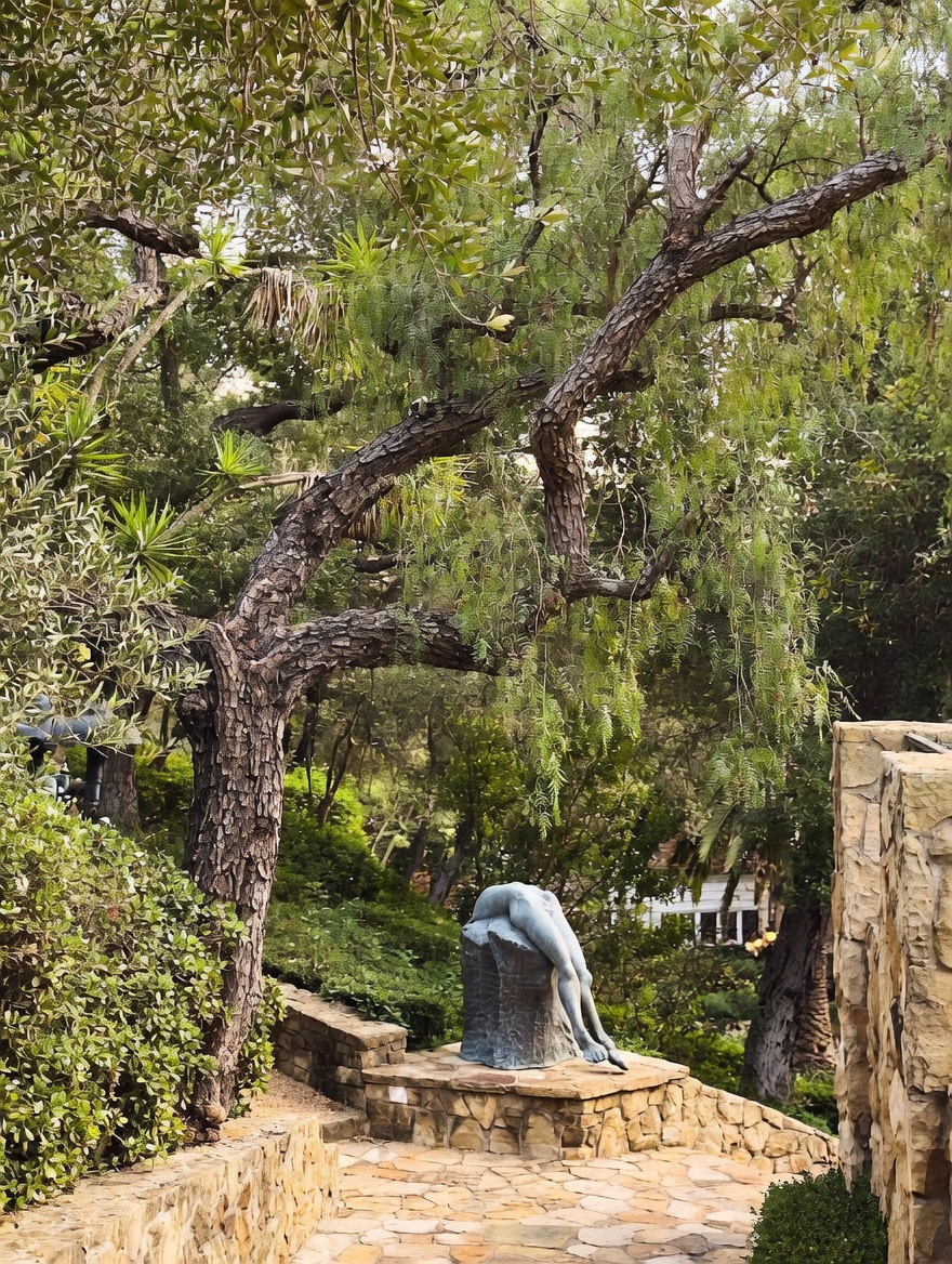 This serene garden setting features a stone-paved patio area highlighted by a unique, sculptural art piece depicting a reclining figure atop a stone pedestal. A large, mature pepper tree with sprawling, textured branches provides a natural canopy over the space, while lush greenery and stone retaining walls create a secluded, tranquil atmosphere. The perspective is a ground-level shot looking toward the sculpture, emphasizing the artistic and peaceful nature of the outdoor living space.