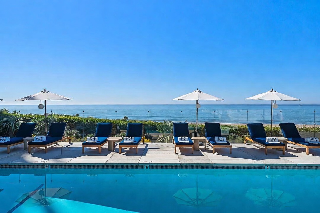 This image captures a serene, sun-drenched swimming pool deck overlooking the ocean. A row of dark blue lounge chairs, each adorned with a white towel, is arranged neatly under white umbrellas, creating a luxurious resort-like atmosphere. The scene is framed by a clear glass railing that provides an unobstructed, panoramic view of the horizon and the calm sea beyond.