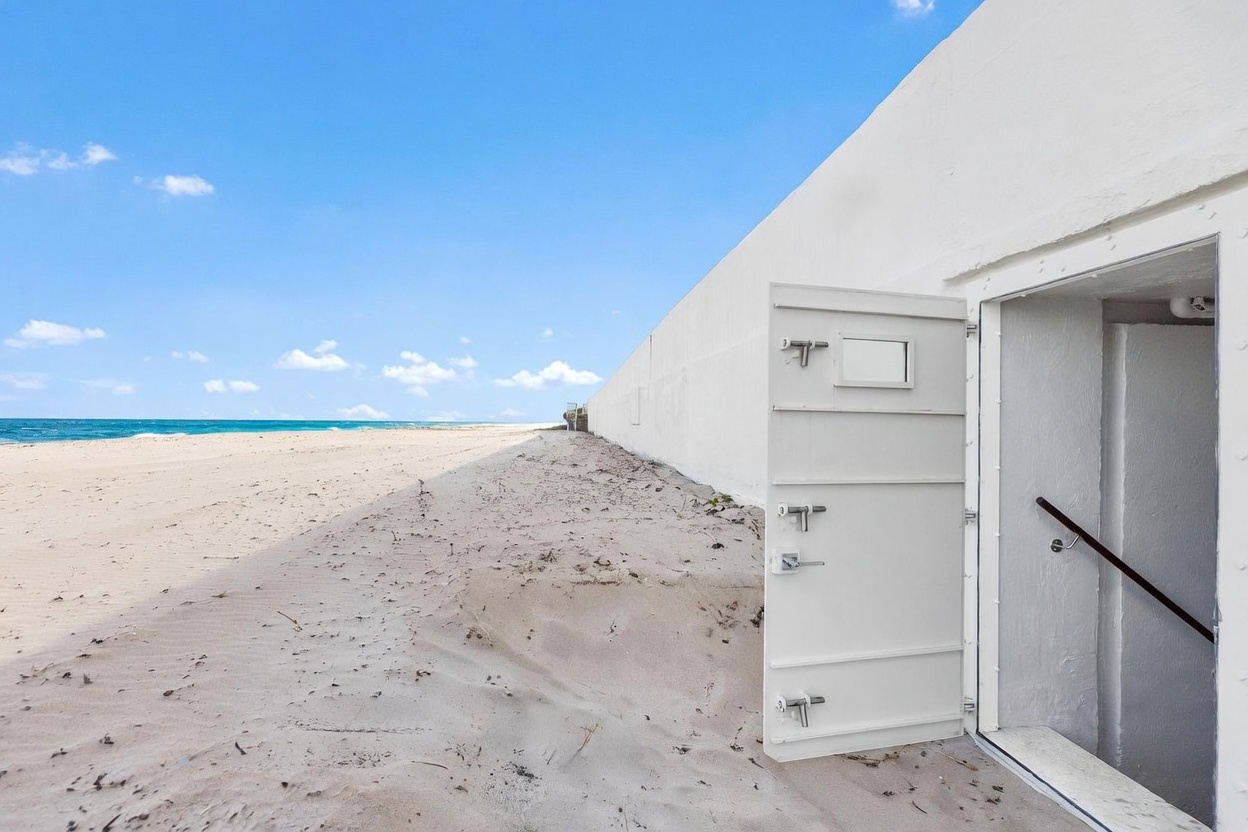This unique exterior view captures a heavy, industrial-style metal door opening directly onto a sandy beach. The door leads into a stairwell, suggesting a subterranean or bunker-style access point integrated into a long, white concrete wall that runs parallel to the shoreline. The bright, clear blue sky and expansive ocean horizon create a stark, minimalist contrast against the utilitarian structure.