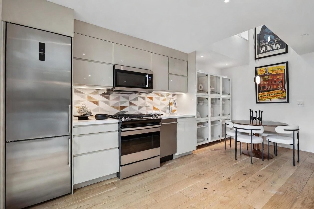 This modern, compact kitchen features sleek, high-gloss cabinetry in a neutral tone paired with stainless steel appliances and a geometric patterned backsplash. A small, circular dining table with four chairs sits adjacent to the kitchen area, accented by framed movie posters on the wall. The space is finished with light wood flooring and clean, contemporary lines, creating a bright and functional living environment.