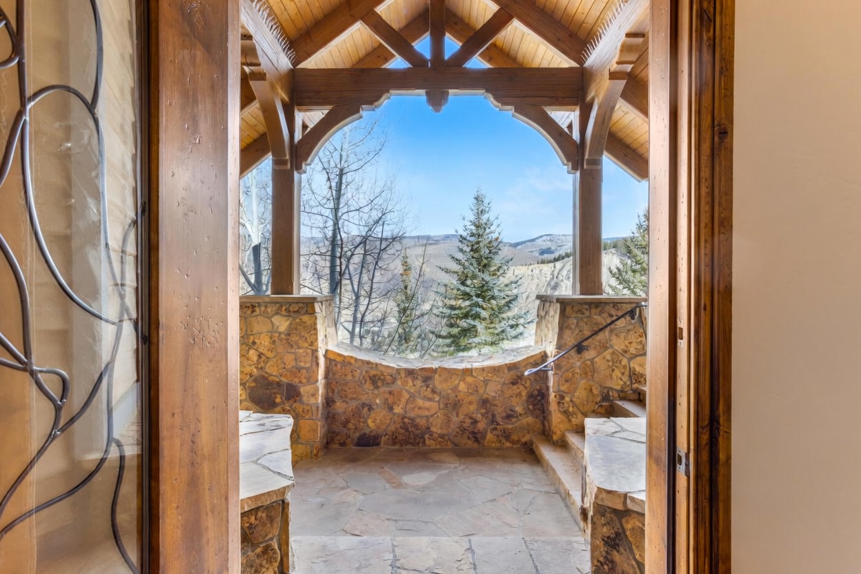 This image captures a view from an interior doorway looking out onto a covered stone patio framed by rustic timber beams. The space features natural stone walls and flooring, with a curved stone railing that overlooks a scenic mountain landscape with evergreen trees. The perspective creates a welcoming transition between the indoor living space and the expansive outdoor scenery.