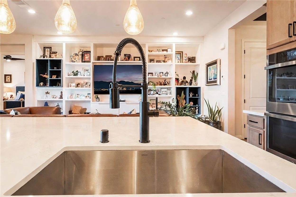 This perspective captures a modern kitchen island with a large stainless steel sink and a dark, high-arc faucet, looking out toward an open-concept living area. The background features a wall of built-in shelving filled with decor and a television, creating a seamless transition between the culinary and relaxation spaces. Warm pendant lighting hangs above the island, contributing to the bright and inviting atmosphere of the home.