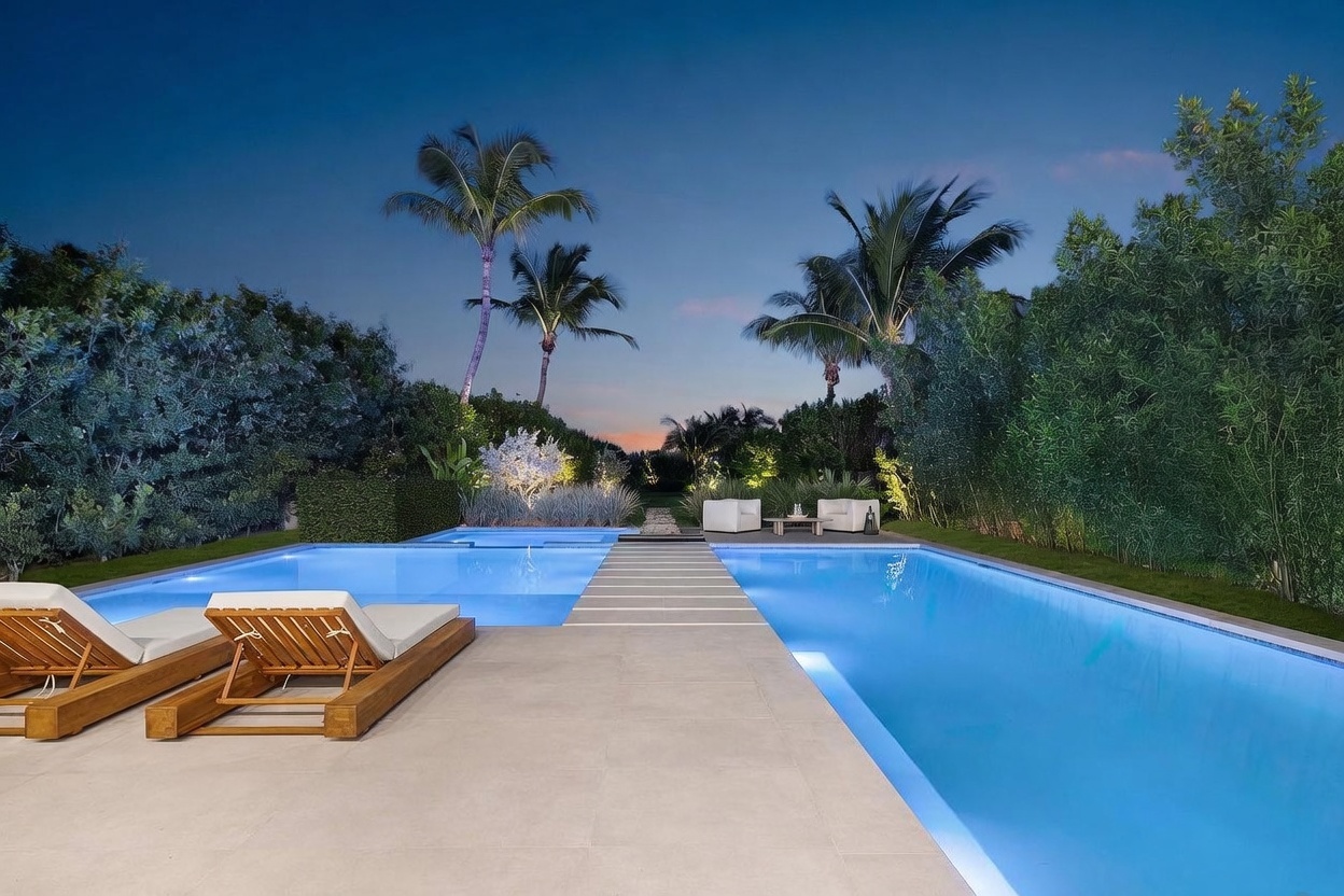 This serene outdoor space features a large, rectangular swimming pool with a central stone walkway bridge that divides the water. Two wooden lounge chairs with white cushions sit on the patio in the foreground, while a pair of modern armchairs and a small table are positioned at the far end of the pool. The scene is framed by lush, dense greenery and tall palm trees, creating a private, resort-like atmosphere during the twilight hour.