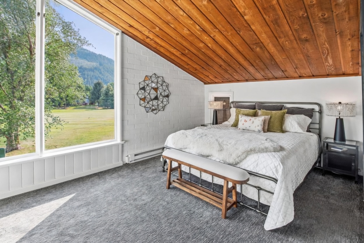 This bedroom features a unique slanted wood-paneled ceiling and a large window that provides an expansive view of the surrounding landscape. The room is furnished with a metal-frame bed topped with a white quilted comforter and a textured throw, complemented by a wooden bench at the foot of the bed. A white painted brick wall serves as an accent, decorated with a geometric metal wall sculpture, creating a rustic yet modern aesthetic.