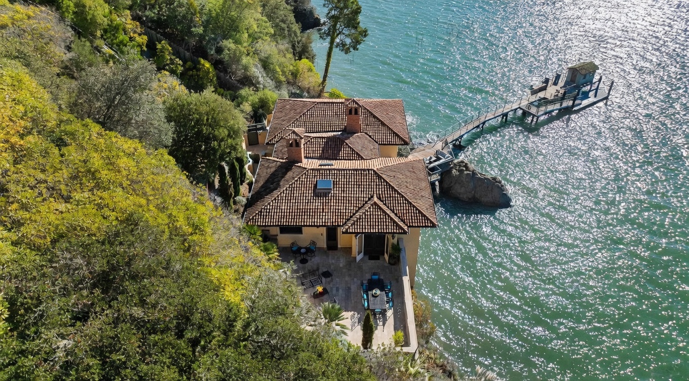 This high-angle aerial view captures a Mediterranean-style villa perched on a rocky cliffside overlooking a sparkling body of water. The home features a multi-tiered terracotta tile roof, a stone-paved patio area with outdoor dining furniture, and a private wooden pier extending into the water. The surrounding landscape is lush with dense trees, creating a secluded and serene waterfront retreat.