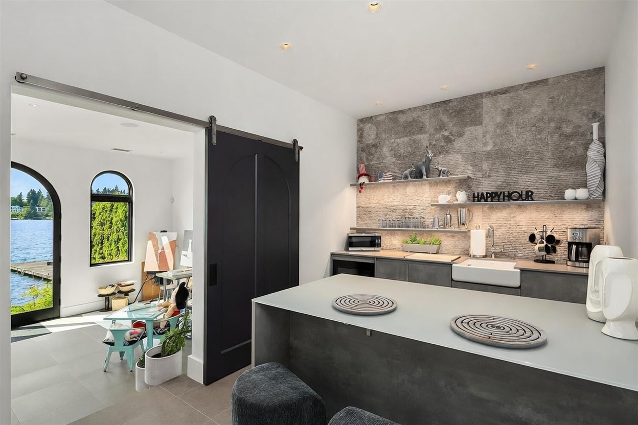 This contemporary kitchen features a sleek island with dark cabinetry and a stone countertop, complemented by a textured stone-clad wall behind the main workspace. Floating shelves and under-cabinet lighting highlight the bar area, which includes a farmhouse-style sink and a coffee station. A large black sliding barn door separates the kitchen from an adjacent sunlit studio space that offers a scenic view of the water through arched windows.