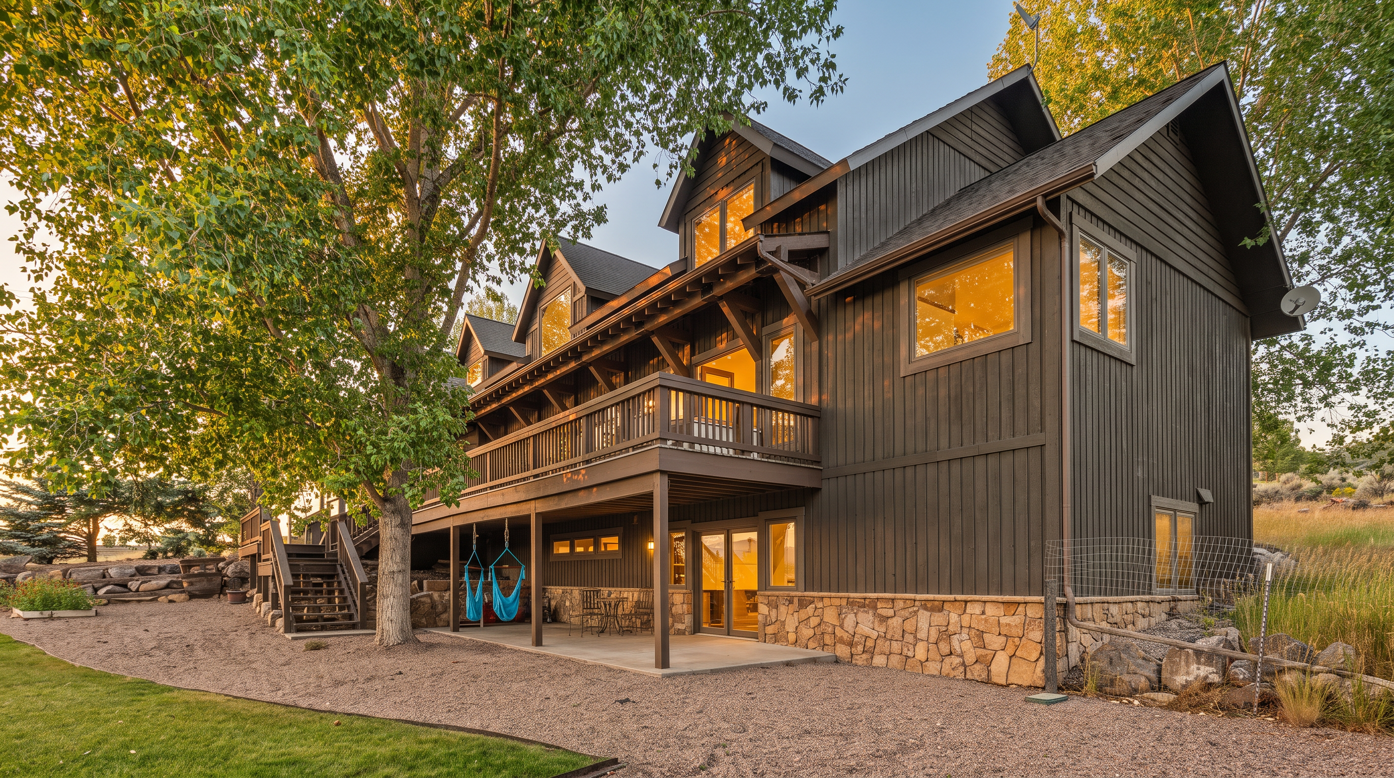 This multi-story mountain-style home features dark vertical cedar siding complemented by a stone foundation and a prominent wrap-around wooden deck. The spacious, shaded backyard features a large gravel patio area ideal for lounging, with comfortable sling chairs under the deck and mature trees providing scenic shade. The overall aesthetic is rustic, cozy, and well-integrated into the natural landscape.