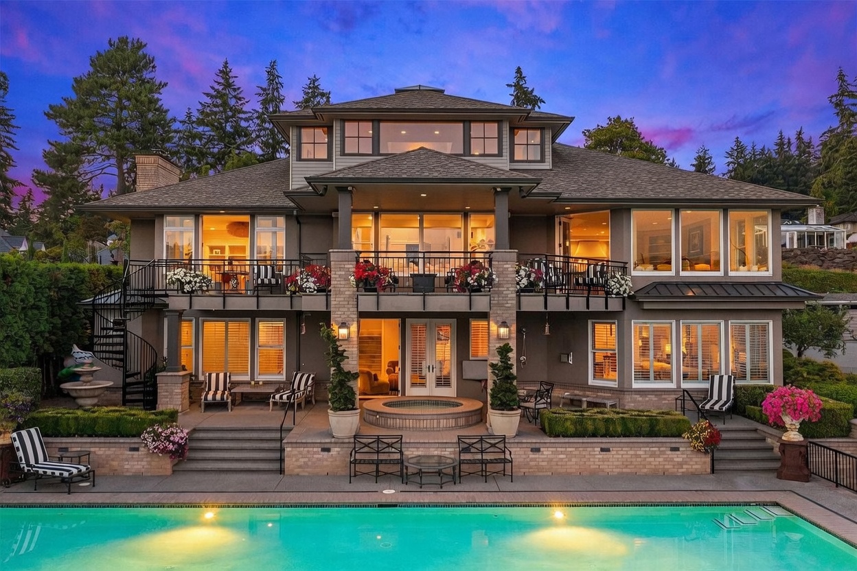 This stunning multi-level residence features a sophisticated rear facade overlooking a private swimming pool and integrated spa. The home showcases a spacious upper-level balcony with black iron railings and a lower-level patio area, both perfect for outdoor entertaining. Large windows and glass doors provide a seamless connection between the interior living spaces and the beautifully landscaped backyard at twilight.