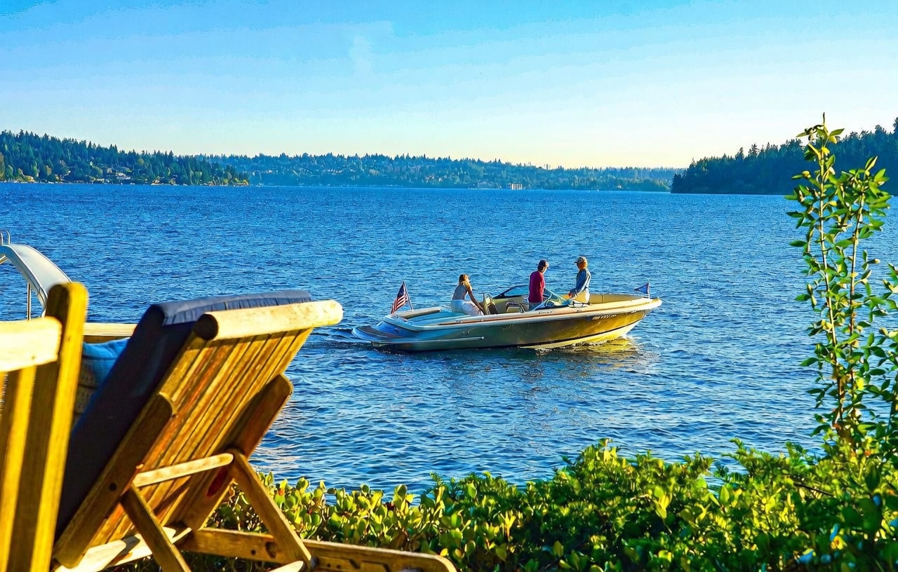 This scenic outdoor view captures a tranquil lakefront setting from a wooden deck, featuring a lounge chair in the foreground and lush greenery. A classic motorboat with three passengers cruises across the calm, deep blue water, framed by a distant, tree-lined shoreline under a clear sky. The perspective emphasizes the peaceful, recreational lifestyle associated with waterfront property living.