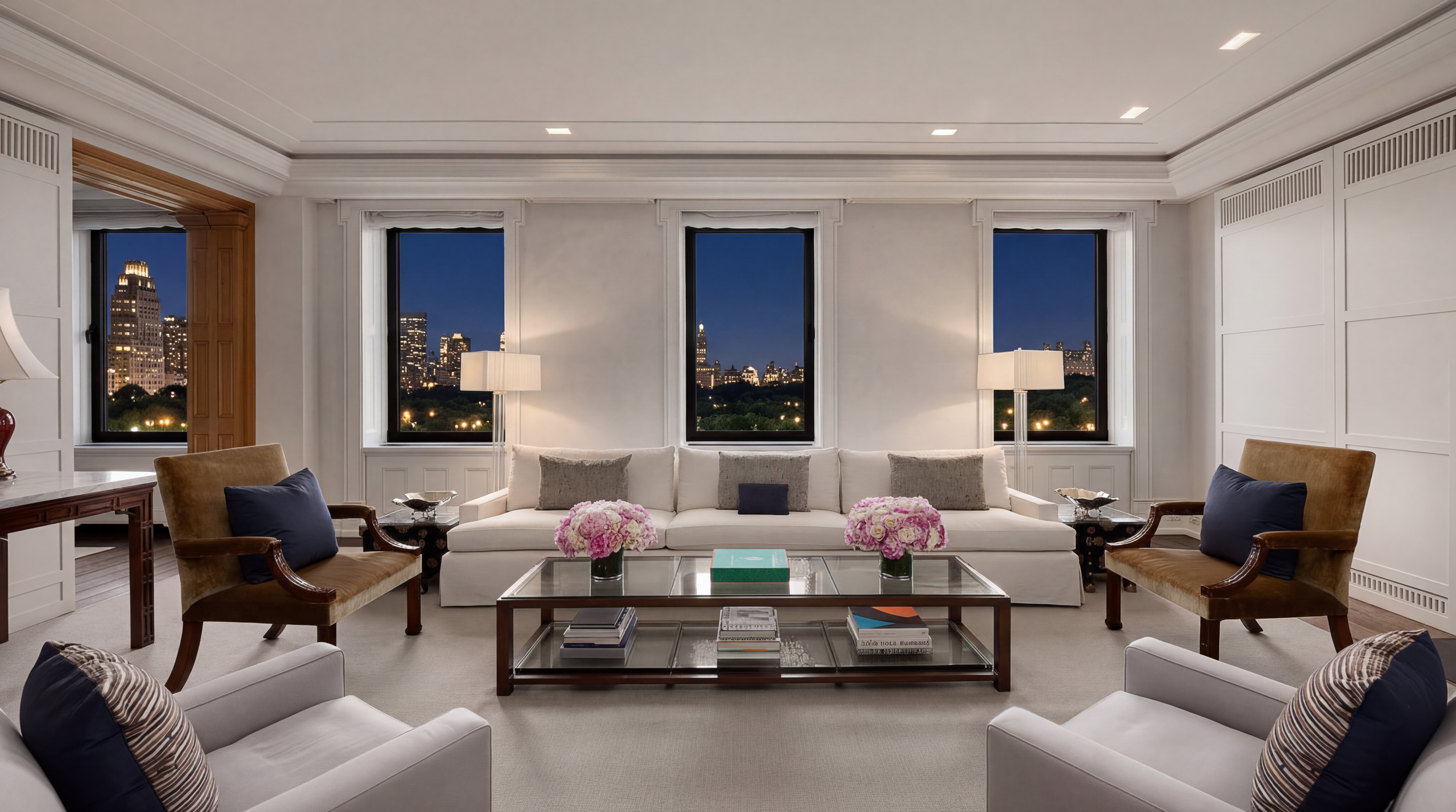 This sophisticated, symmetrical living room features a generous cream-colored sofa flanked by two floor lamps, set against a backdrop of three large windows providing stunning evening views of a city skyline and park. The room is elegantly furnished with caramel velvet armchairs, light grey lounge chairs, and a central glass-topped coffee table adorned with floral arrangements and books. Its pristine walls, refined millwork, and balanced layout create a luxurious and inviting atmosphere perfect for high-end urban living.