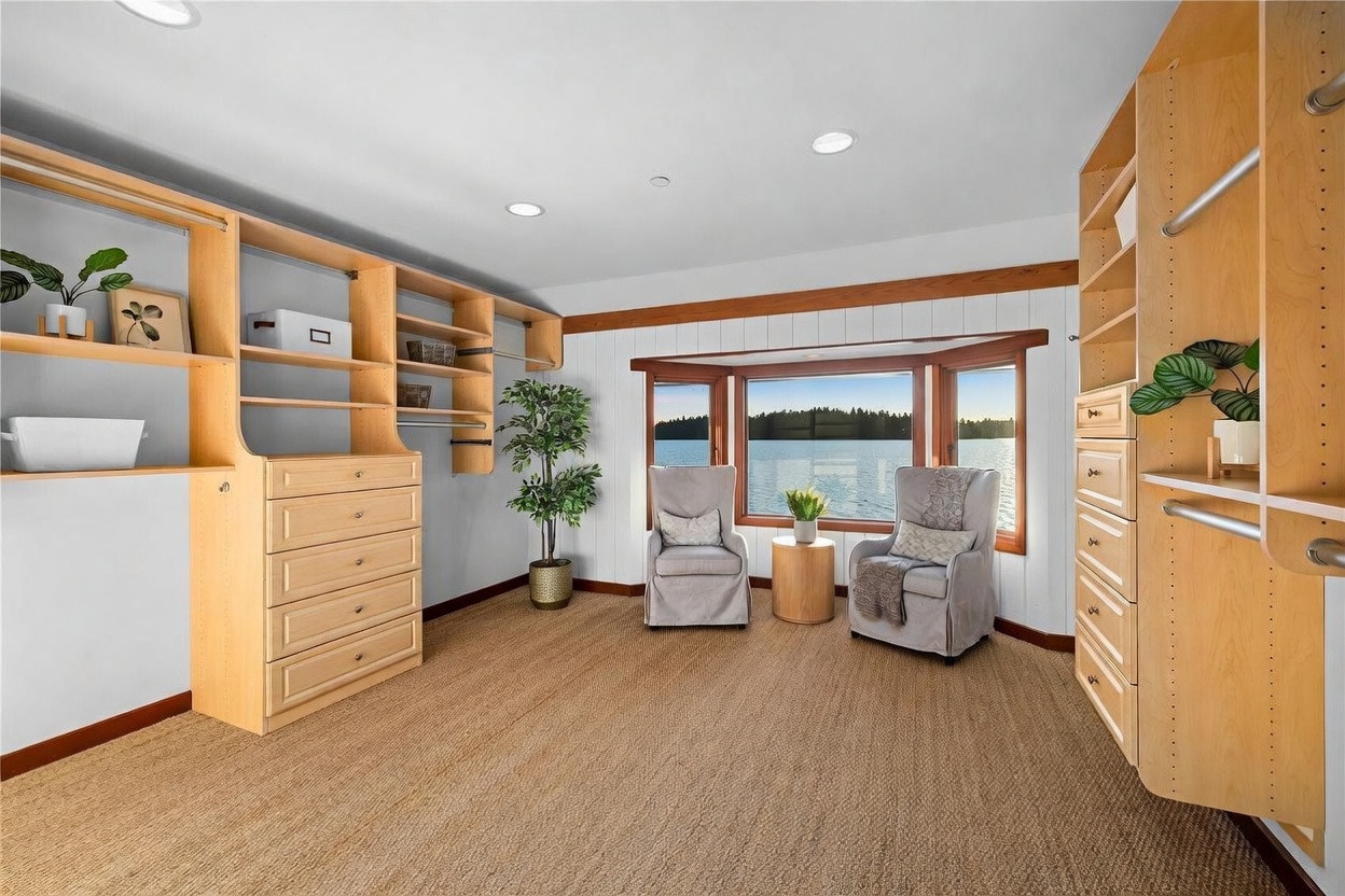 This spacious walk-in closet features extensive light-wood custom shelving and built-in cabinetry, providing ample storage and organization. A cozy seating area with two armchairs and a small side table is positioned in front of a large bay window, offering a serene, panoramic view of the water. The room is finished with neutral carpeting and recessed lighting, creating a bright and tranquil atmosphere.