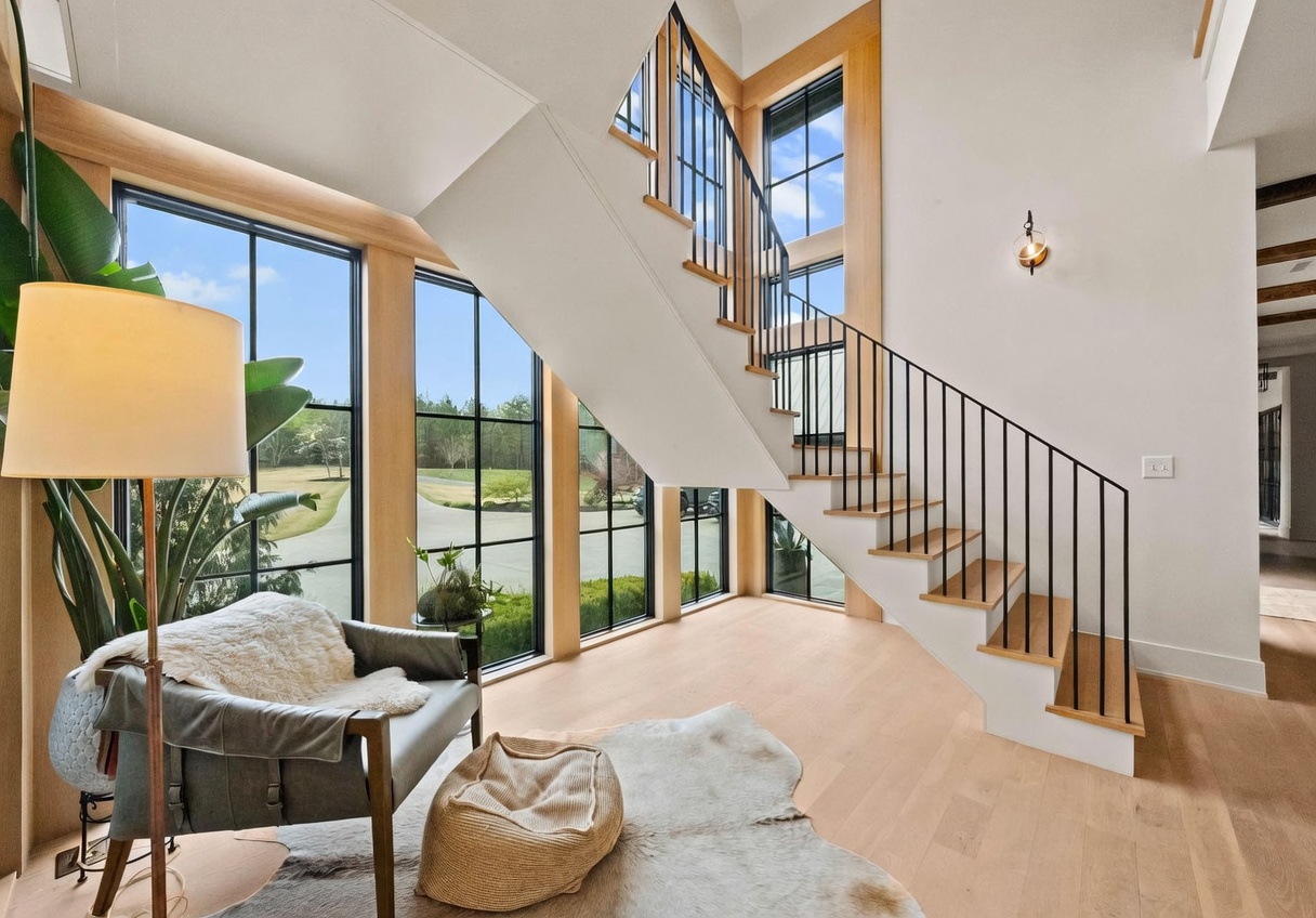 This bright, modern entryway features a striking staircase with light wood treads and a minimalist black metal railing. Large, multi-pane windows framed in light wood provide an expansive view of the outdoors, while a cozy seating area with a leather chair and a textured rug creates a welcoming nook beneath the stairs. The space is characterized by clean white walls, light wood flooring, and a warm, airy aesthetic.