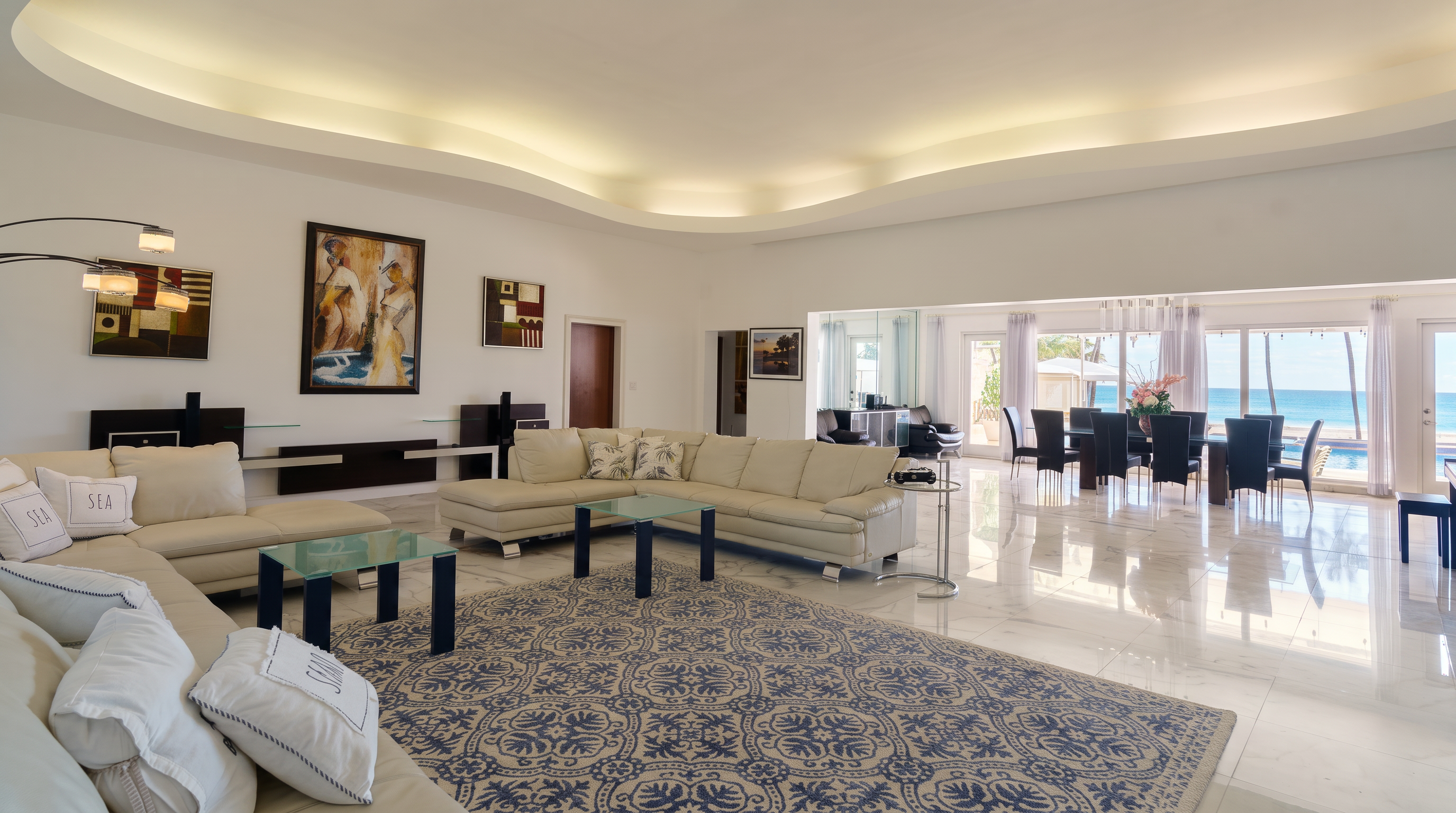 This expansive, open-concept living area features a large, light-colored sectional sofa arranged on a patterned area rug over polished stone flooring. The room is characterized by a curved, recessed ceiling with integrated lighting and a seamless transition to a dining area that overlooks the ocean through large glass doors. The space is bright and airy, decorated with contemporary artwork and minimalist furniture, creating a sophisticated and inviting atmosphere.