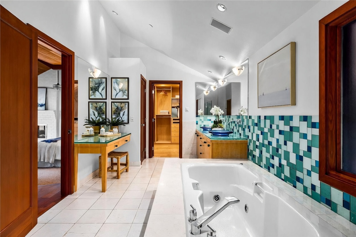 This spacious primary bathroom features a luxurious jetted soaking tub set against a vibrant, multi-toned blue mosaic tile wall. The room includes a dedicated vanity area with a glass countertop, a walk-in closet visible through an open doorway, and a double vanity with blue glass sinks. The high, vaulted ceiling and neutral tile flooring create an airy, sophisticated atmosphere with a touch of mid-century modern flair.