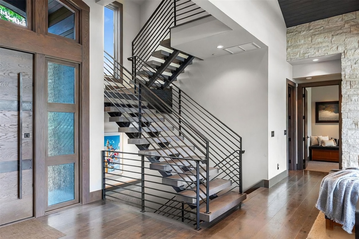 This grand entryway features a striking open-riser staircase with dark metal railings and thick wood treads, serving as the focal point of the space. To the left, a large, multi-pane wood-framed door allows natural light to flood the area, while a stone-clad wall on the right adds texture and depth. The perspective captures the transition from the entryway into a secondary hallway, showcasing the high-end, modern architectural design and rich wood flooring.