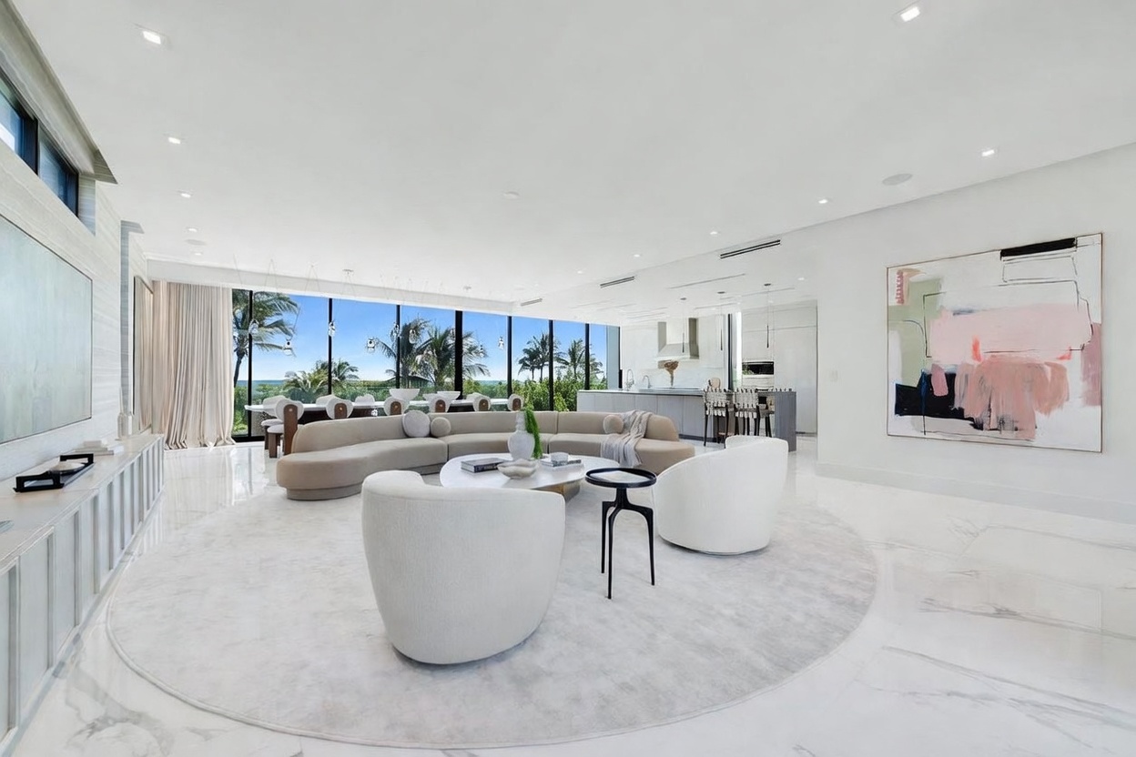 This expansive, open-concept living area features a large, curved sectional sofa arranged on a circular area rug, creating a sophisticated conversation space. The room is bathed in natural light from floor-to-ceiling windows that offer a panoramic view of the outdoors, while a large abstract painting adds a vibrant focal point to the minimalist white walls. The space flows seamlessly into a modern kitchen area, emphasizing a bright, airy, and luxurious aesthetic.