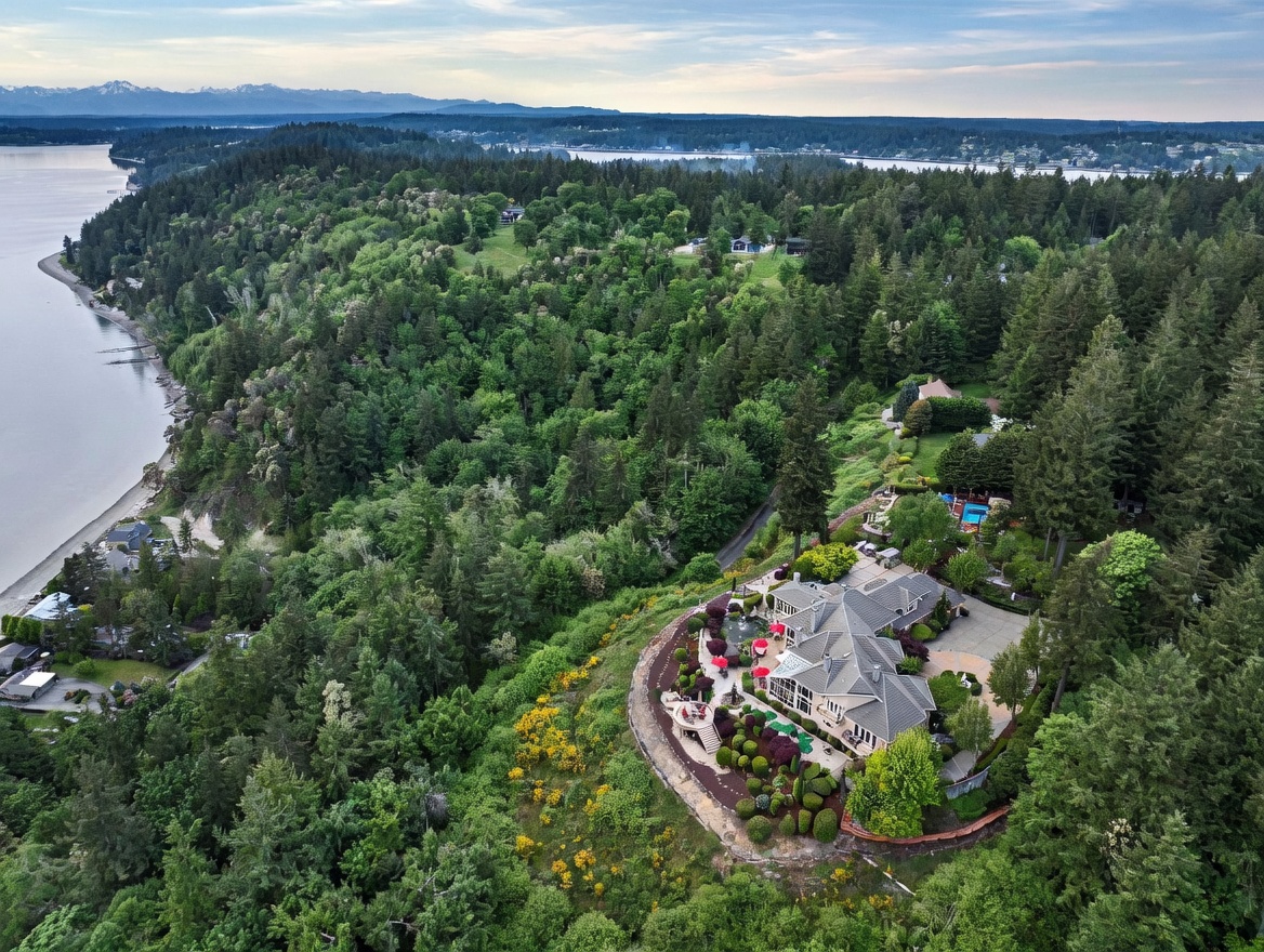 This high-angle aerial view captures a sprawling luxury estate perched on a forested cliffside overlooking a vast body of water. The property features a multi-gabled residence with extensive landscaping, a tiered patio area, and a private driveway, all surrounded by dense evergreen trees. The cinematic perspective emphasizes the home's secluded, elevated position and its dramatic connection to the natural coastal landscape.