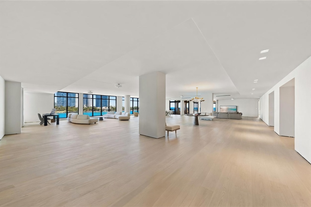 This expansive, open-concept living area features light wood flooring and a minimalist aesthetic, emphasizing a sense of vast space. Large windows line the far wall, providing panoramic views of the water and city skyline, while a central structural pillar anchors the room. The space is sparsely furnished with contemporary seating arrangements, creating a bright, airy, and sophisticated atmosphere.