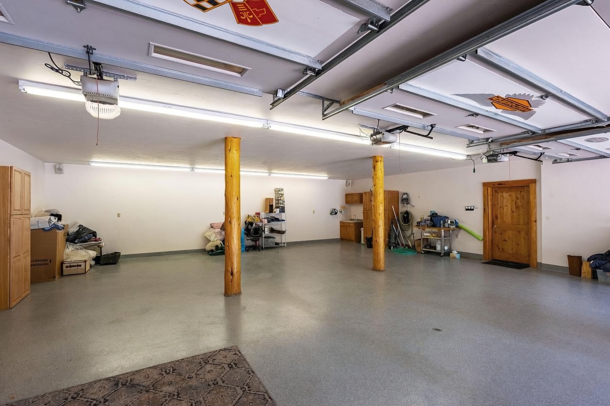 This spacious, multi-car garage features a clean, epoxy-coated floor and two prominent, rustic log support columns that anchor the center of the room. The ceiling is equipped with overhead garage door tracks and bright, linear fluorescent lighting, while a small kitchenette area with wooden cabinetry is tucked against the back wall. A wooden door provides access to the interior, and the space is decorated with overhead automotive-themed signage, creating a functional and organized workshop or storage environment.