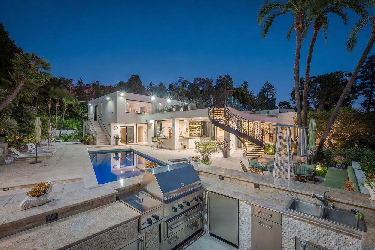 This evening shot captures a luxurious outdoor living space featuring a rectangular swimming pool and a fully equipped stone-clad outdoor kitchen in the foreground. A striking spiral staircase leads to an upper-level terrace, while the modern home architecture is illuminated by warm accent lighting. The scene is framed by tall palm trees and lush landscaping, creating an inviting and sophisticated atmosphere for outdoor entertaining.