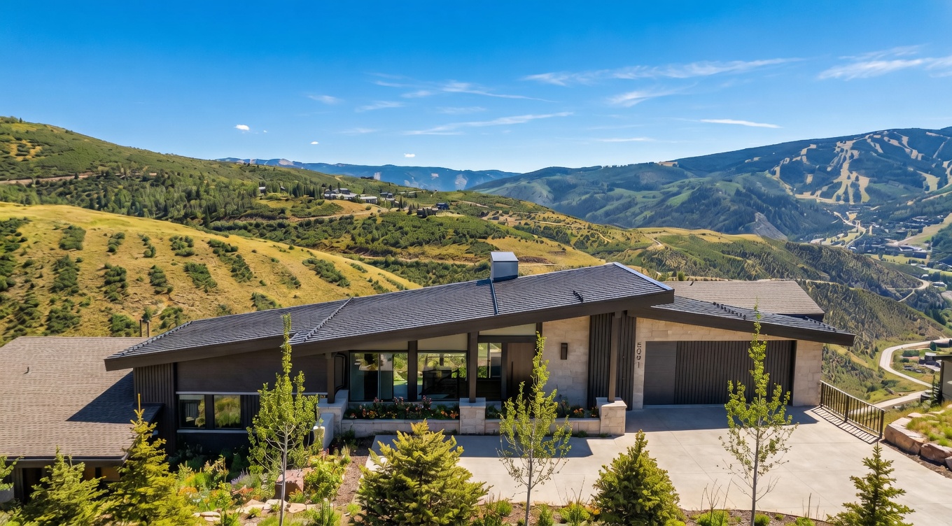 This is a front exterior view of a modern home nestled in a mountainous landscape. The house features a unique roofline, a combination of stone and dark wood siding, and large windows that offer stunning views. The property is surrounded by natural terrain with sparse vegetation and snow patches, creating a serene and private setting.