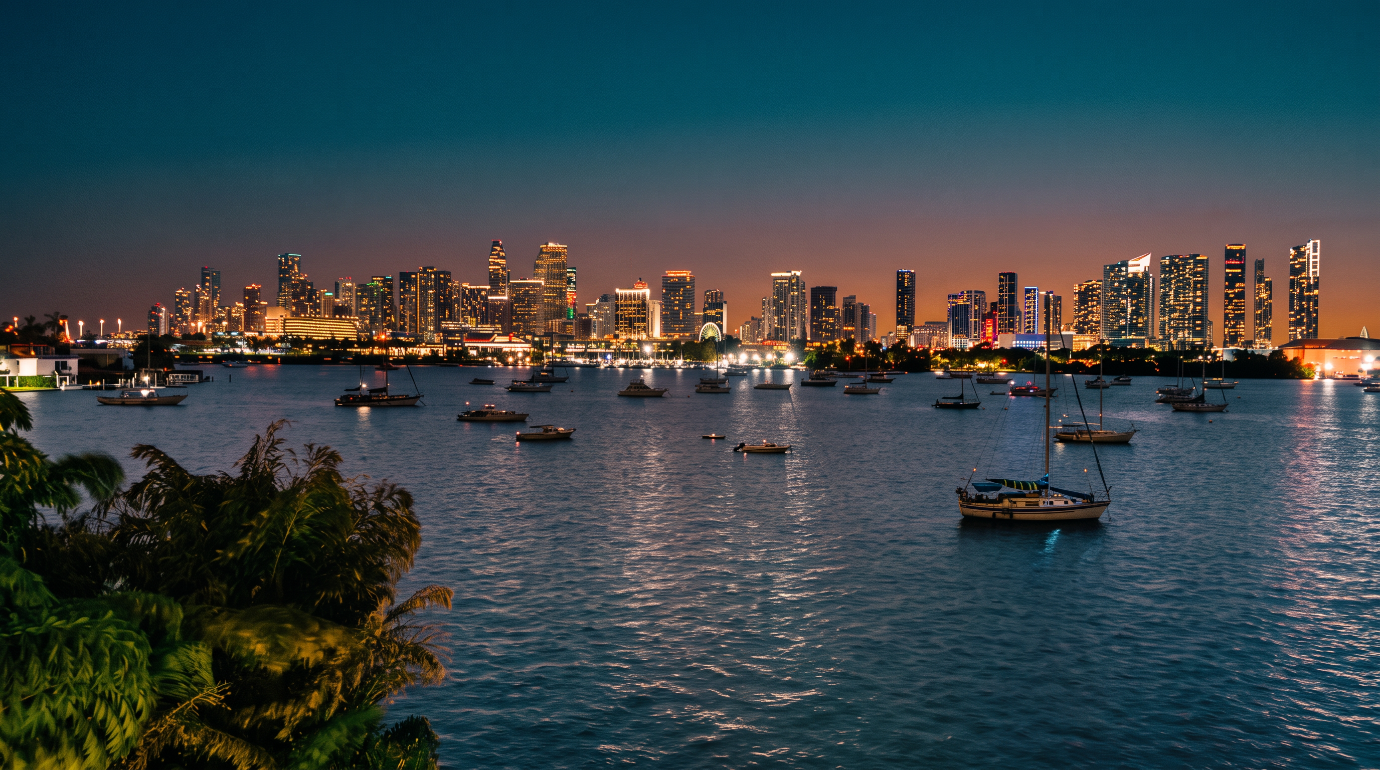 This image captures a scenic, wide-angle view of a city skyline at dusk, framed by lush foreground foliage and a calm body of water filled with anchored sailboats. The perspective is elevated, likely from a balcony or high-rise window, showcasing the vibrant urban lights reflecting across the bay. The composition emphasizes the contrast between the natural elements in the foreground and the dense, illuminated architecture of the metropolitan skyline in the distance.