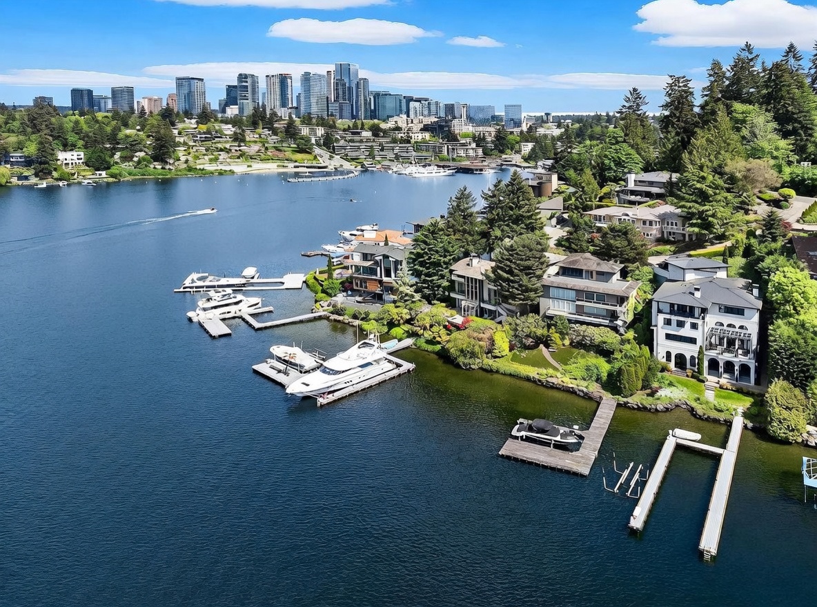 This high-angle aerial view captures a luxurious waterfront property situated on a serene lake, featuring multiple private docks and a large yacht moored at the shore. The multi-story residences are nestled among lush, mature trees, offering a private and tranquil setting with a clear view of the distant city skyline across the water. The composition emphasizes the seamless integration of high-end residential living with direct water access and expansive outdoor spaces.