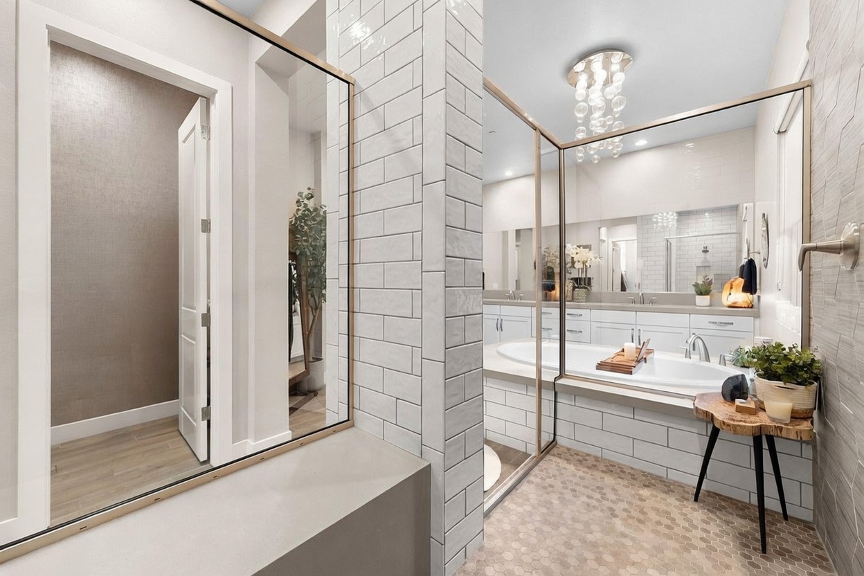 This modern primary bathroom features a deep soaking tub positioned against a wall with white subway tile and a large mirror. A unique, tall, narrow mirror panel separates the vanity area from the entryway, while a small wooden side table with a plant adds a natural touch to the space. The room is finished with patterned floor tiles and a contemporary chandelier, creating a bright and luxurious atmosphere.