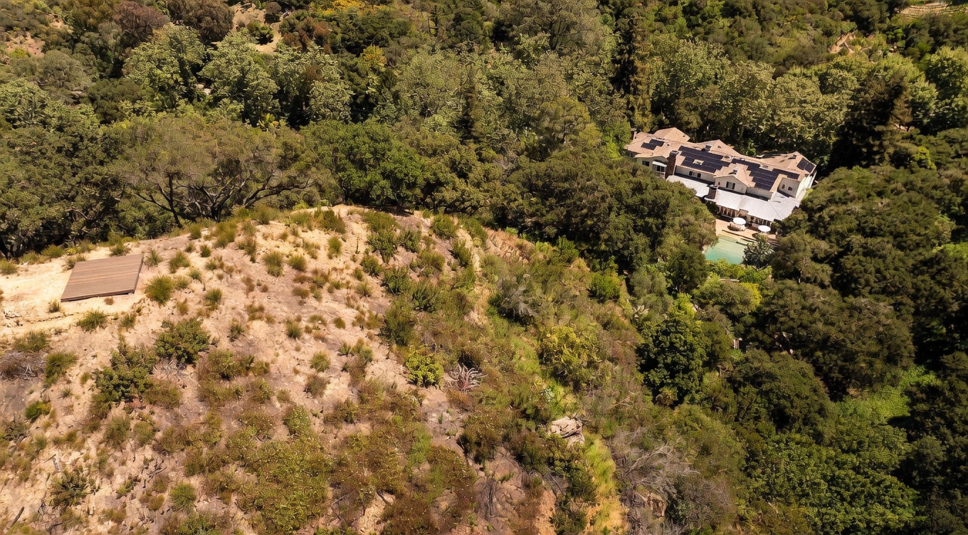 This high-angle aerial view captures a sprawling estate nestled within a lush, wooded hillside. The property features a large multi-level residence with a swimming pool, situated adjacent to a prominent, dry, grassy hillside that includes a flat, elevated wooden platform. The perspective emphasizes the secluded, natural setting and the expansive scale of the grounds.
