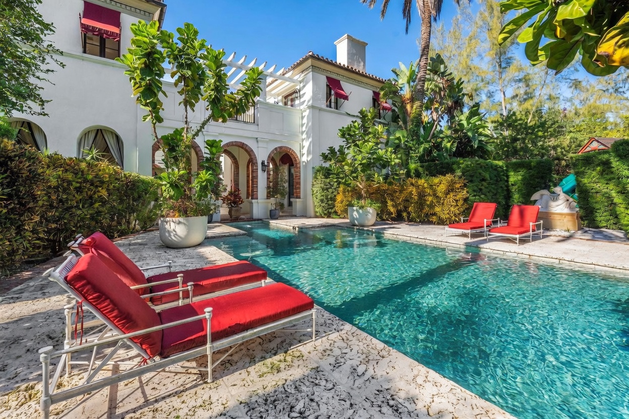 This elegant Mediterranean-style home features a private swimming pool set against a white stucco facade with arched doorways and red awnings. The pool deck is paved with light-colored stone, complemented by vibrant red lounge chairs and lush tropical landscaping, including tall potted fiddle leaf fig trees. The scene captures a serene, resort-like atmosphere perfect for outdoor relaxation and entertaining.