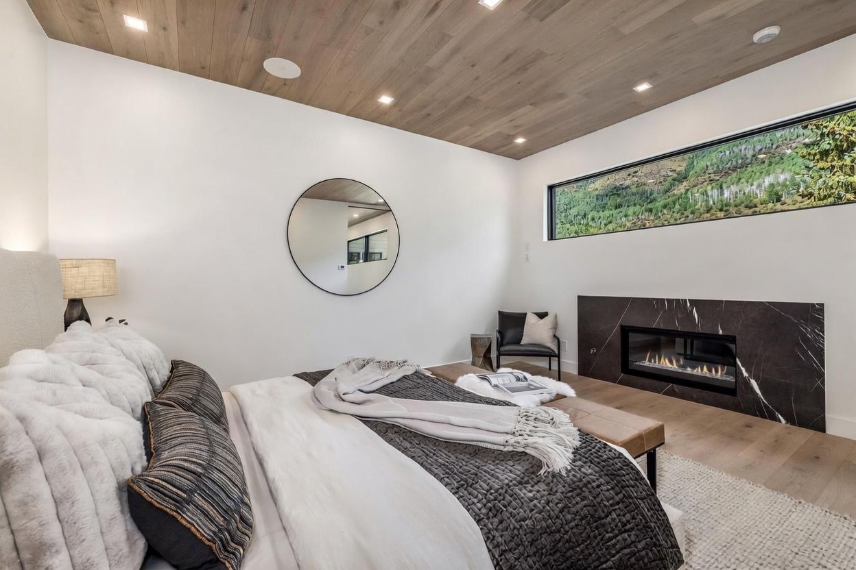 This modern primary bedroom features a warm wood-paneled ceiling and a striking dark stone fireplace surround that anchors the room. A large, horizontal window provides a scenic view of the outdoors, while a circular mirror adds a contemporary design element to the white walls. The space is furnished with a plush bed, a leather bench at the foot, and a sleek accent chair, creating a sophisticated and tranquil atmosphere.