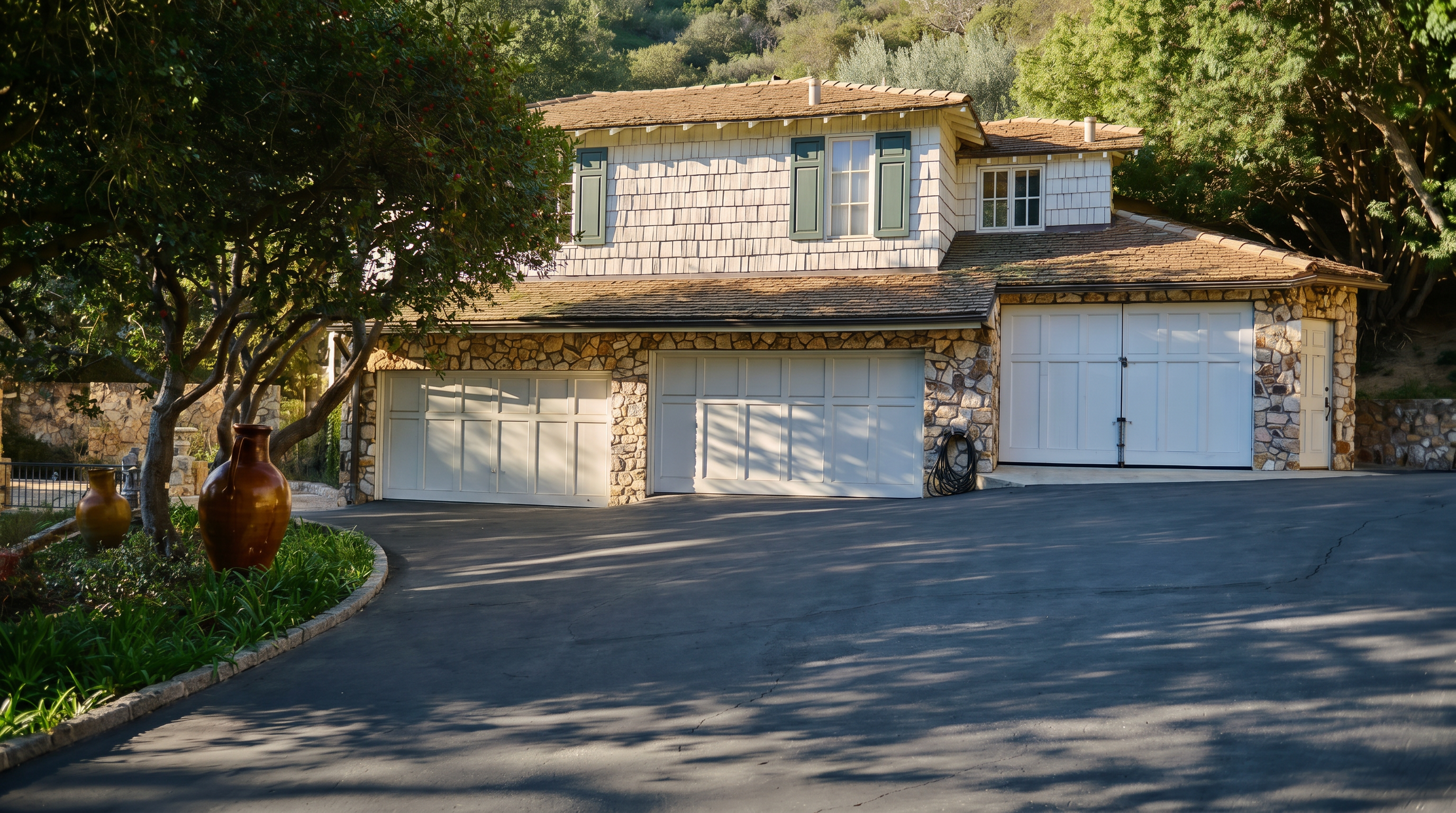 This front-facing view showcases a charming two-story home with a distinctive stone facade and white shingle siding. The property features a spacious asphalt driveway leading to a multi-car garage, surrounded by lush greenery and mature trees. The overall aesthetic is rustic and inviting, set against a backdrop of a gentle hillside.