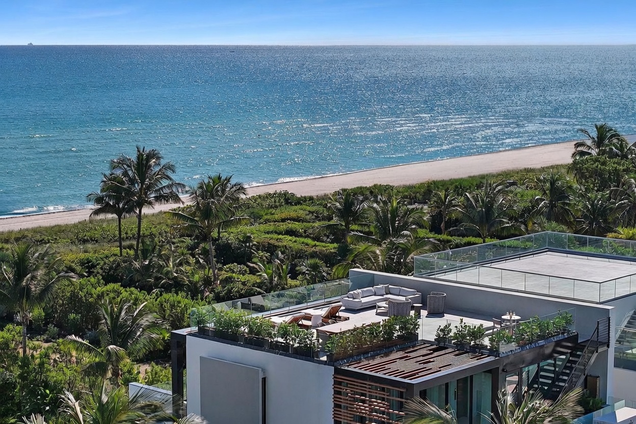This high-angle aerial shot captures a modern luxury residence featuring a spacious rooftop terrace equipped with contemporary outdoor lounge furniture. The property is nestled within lush tropical landscaping, providing a seamless transition to the adjacent sandy beach and the expansive blue ocean beyond. The architectural design emphasizes clean lines and open-air living, perfectly positioned to take advantage of the coastal scenery.