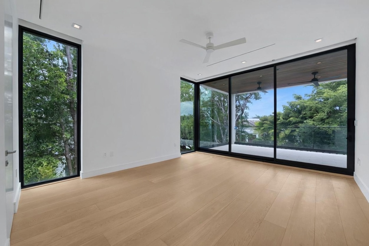This bright and airy room features light wood flooring and crisp white walls, creating a minimalist and modern aesthetic. The space is defined by expansive floor-to-ceiling windows and sliding glass doors that provide abundant natural light and a seamless transition to an outdoor balcony. A ceiling fan is centered in the room, which overlooks a lush, tree-filled exterior landscape.